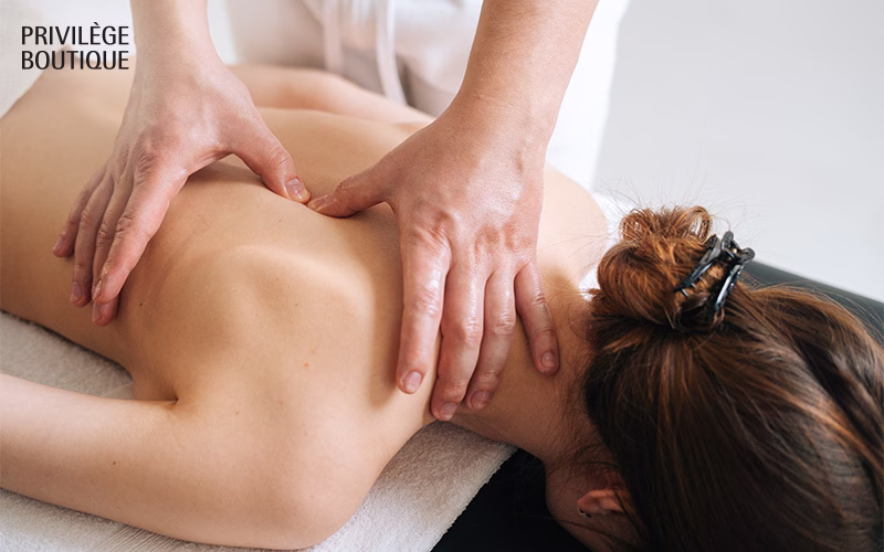 Person lying face-down on a massage table while a massage therapist applies pressure to their upper back and shoulders.