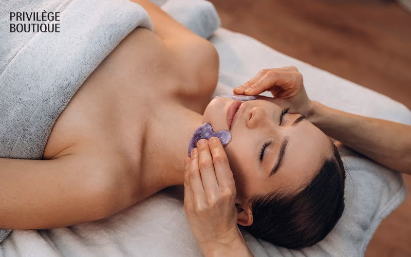 Person receiving facial massage with purple gua sha tool in spa-like setting, lying relaxed on spa bed
