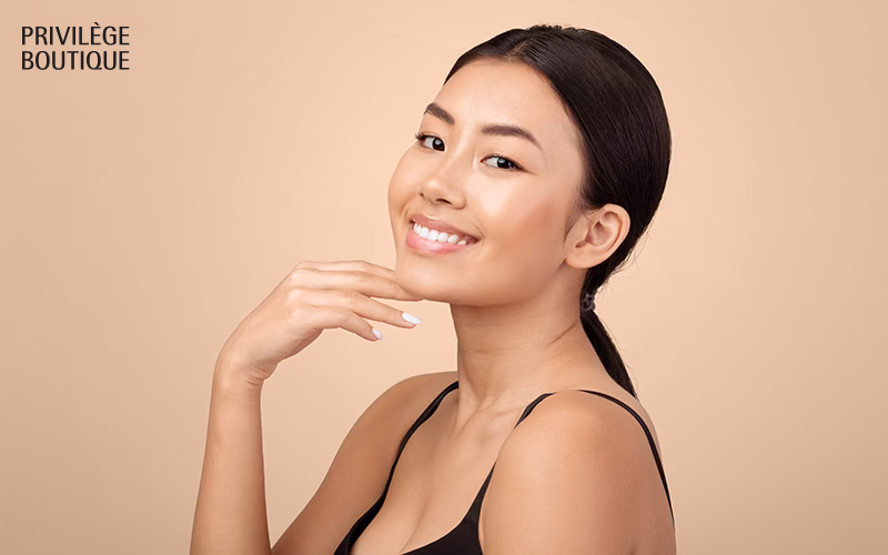 Smiling woman with glowing skin gently touching her face against a light blue background.